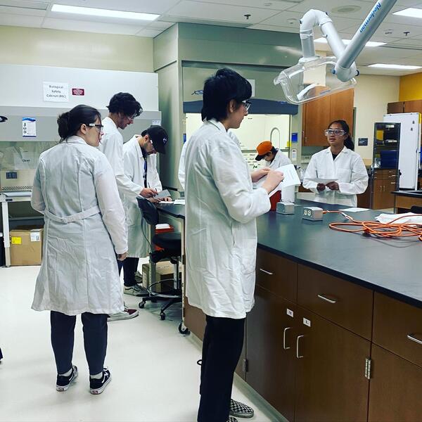 students in white lab coats gathered around a lab bench